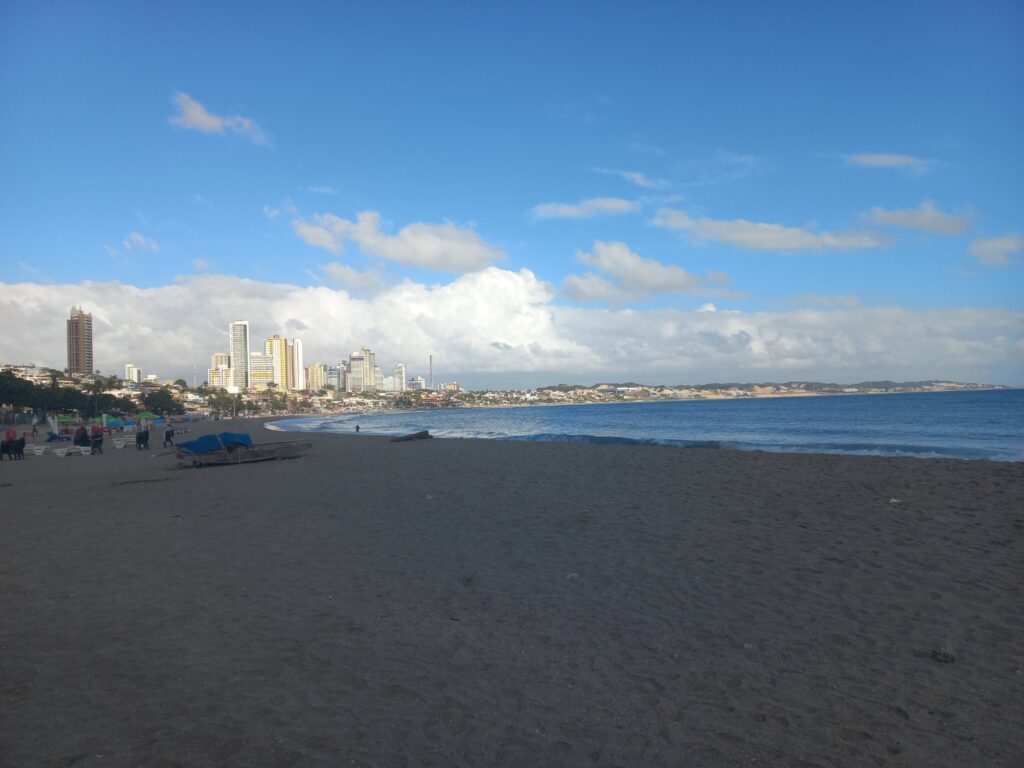 Areia da praia de Ponta Negra pela manhã, com o mar, um barco e os prédios da orla ao fundo