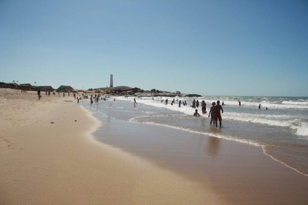 Faixa de areia da Praia da Pedra do Sal com banhistas e o farol ao fundo