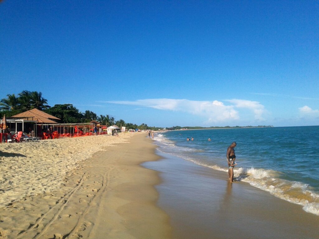 Faixa de areia da Praia de Taperapuã com barracas e guarda-sóis à esquerda e mar calmo
