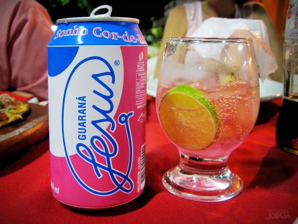 Lata de Guaraná Jesus ao lado de um copo com o refrigerante cor-de-rosa, gelo e limão