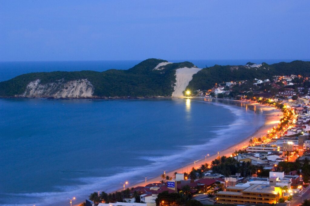 Orla de Ponta Negra ao entardecer, com as luzes dos hotéis acesas e o Morro do Careca ao fundo
