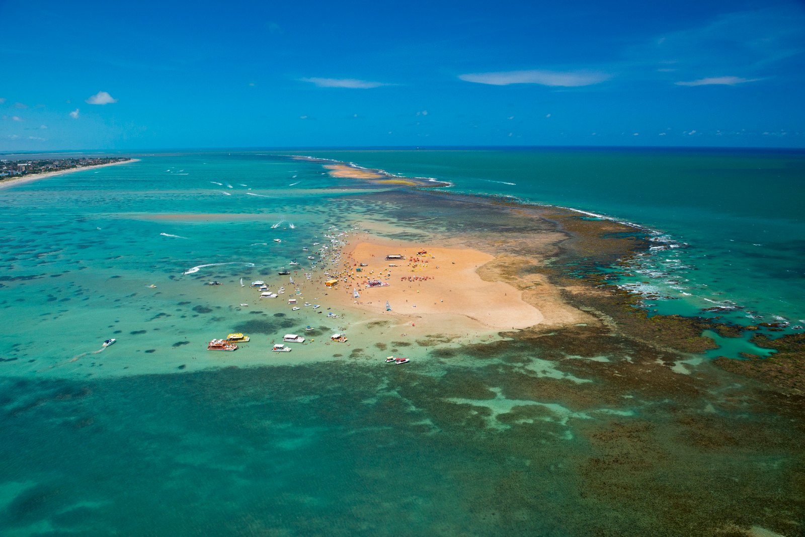 Vista aérea do banco de areia de Areia Vermelha, em Cabedelo, exposto na maré baixa, cercado de recifes e mar verde-claro, com barcos ancorados