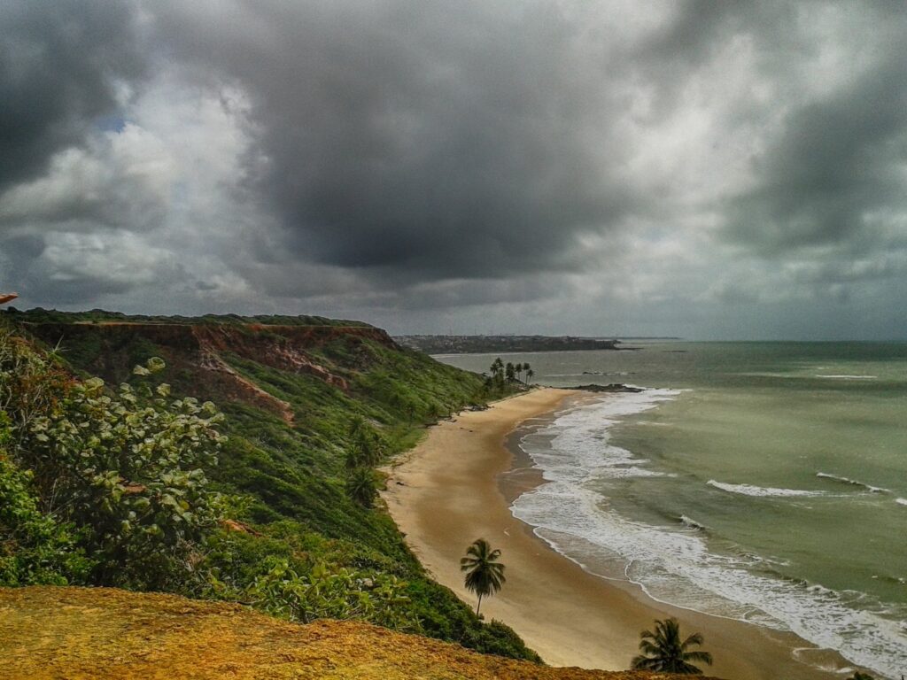 Praia de Coqueirinho vista do alto da falésia sob céu carregado de nuvens, com a faixa de areia e o mar esverdeado