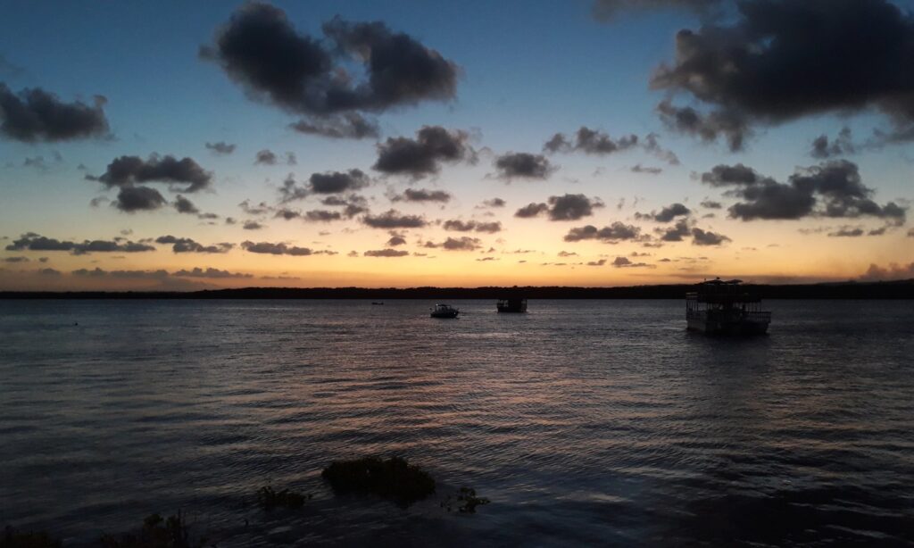 Entardecer sobre o rio Paraíba visto da Praia do Jacaré, em Cabedelo, com barcos de passeio na água e nuvens escuras no céu alaranjado