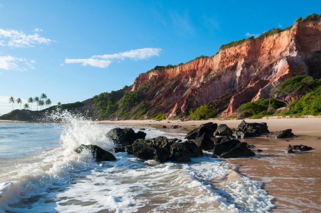 Falésias coloridas da Praia de Tabatinga, no Conde, com pedras escuras na areia, ondas quebrando e coqueiros ao fundo sob céu azul