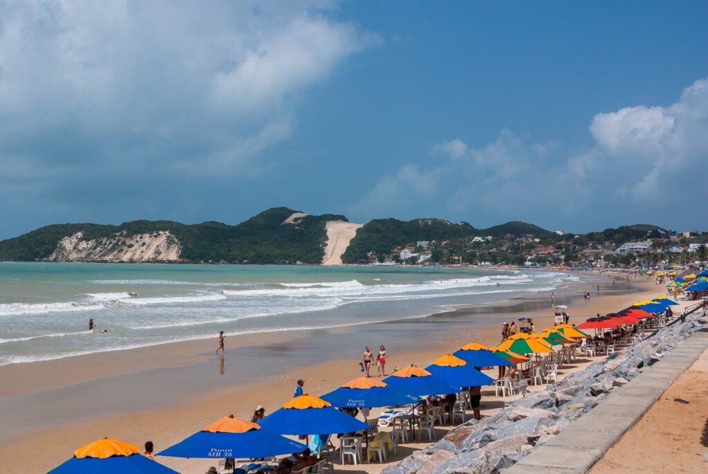 Praia de Ponta Negra em dia de sol, com guarda-sóis coloridos na areia, mar calmo e o Morro do Careca ao fundo