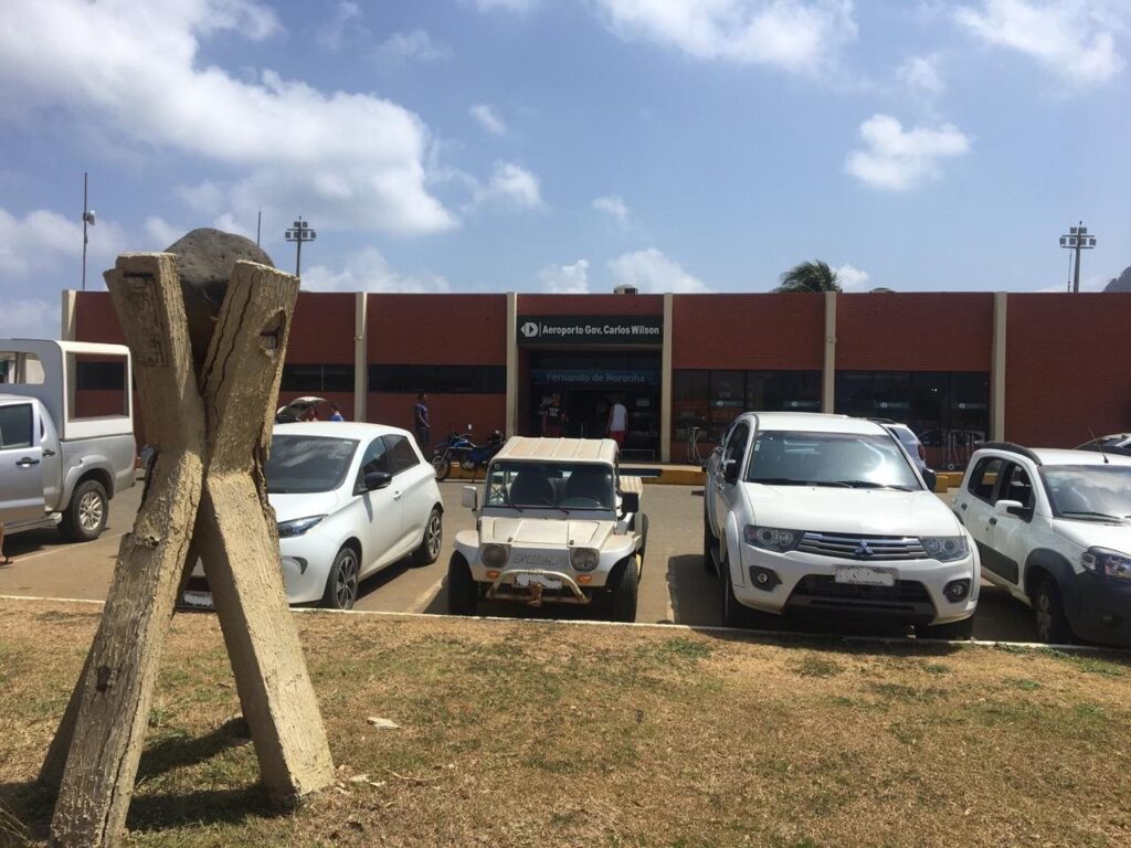 Fachada do aeroporto de Fernando de Noronha com carros e buggies estacionados em frente, sob céu com nuvens