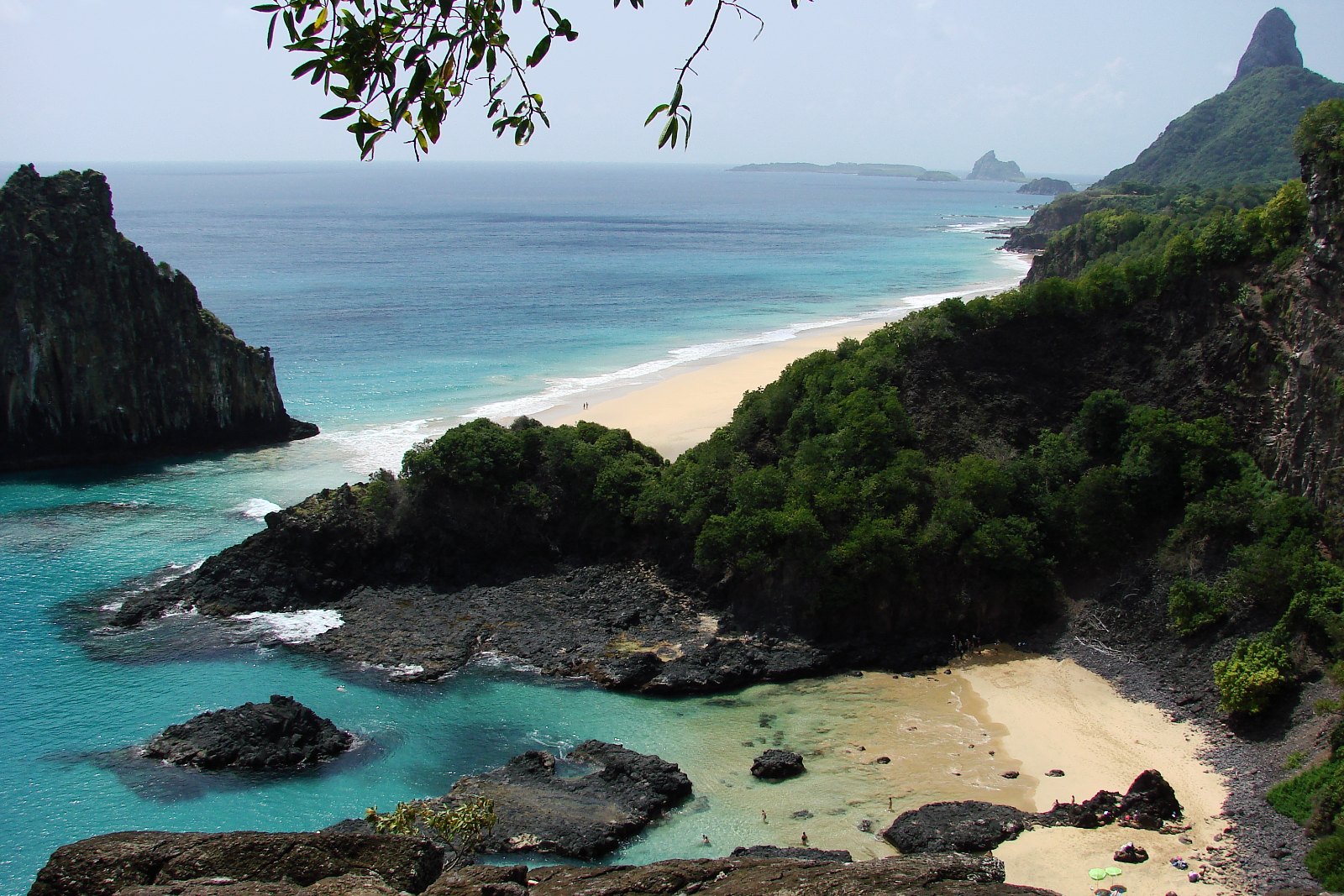 Baía dos Porcos com piscinas de pedra, uma longa faixa de areia ao fundo, o Morro Dois Irmãos à esquerda e o Morro do Pico no horizonte