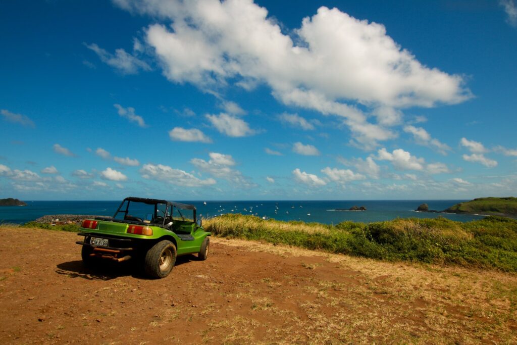 Buggy verde estacionado em um mirante de terra vermelha, com o mar e o céu azul de Fernando de Noronha ao fundo