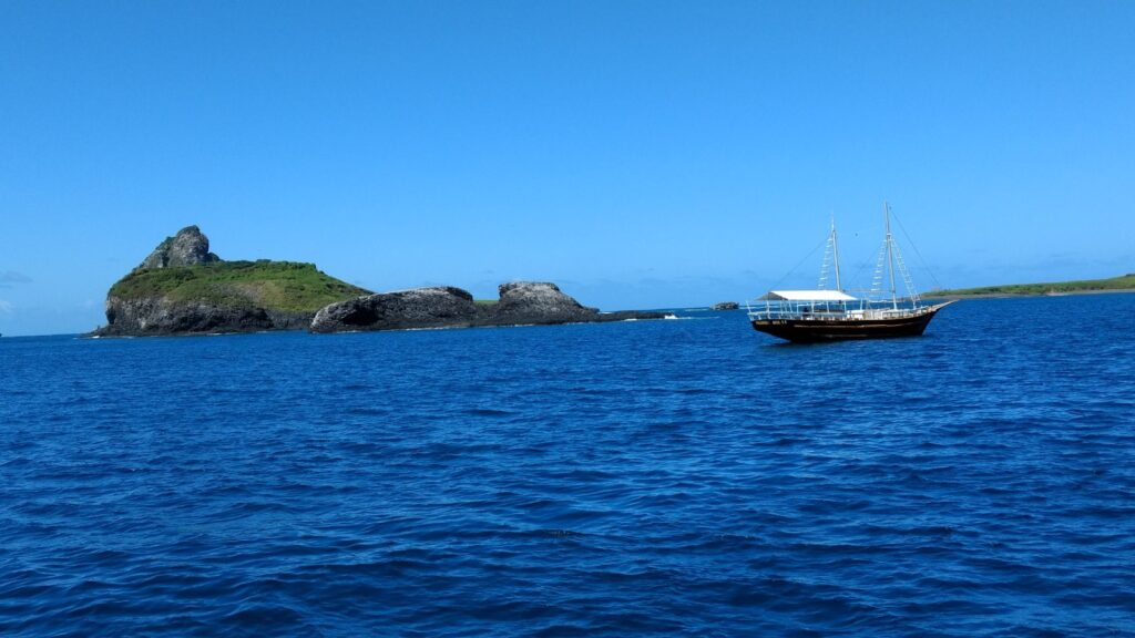 Veleiro de passeio navegando em mar azul-escuro perto de ilhotas rochosas de Fernando de Noronha