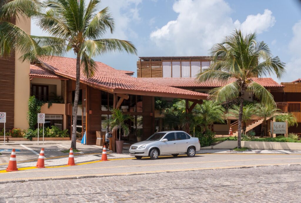 Entrada de hotel com telhado de cerâmica e coqueiros em rua de paralelepípedos de Ponta Negra