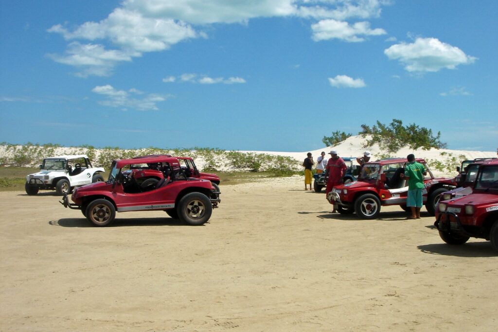 Buggies vermelhos estacionados na areia ao lado de uma duna, com turistas, em Jericoacoara