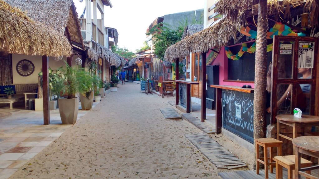 Rua de areia da vila de Jericoacoara com restaurantes e lojas de telhado de palha e mesas na calçada