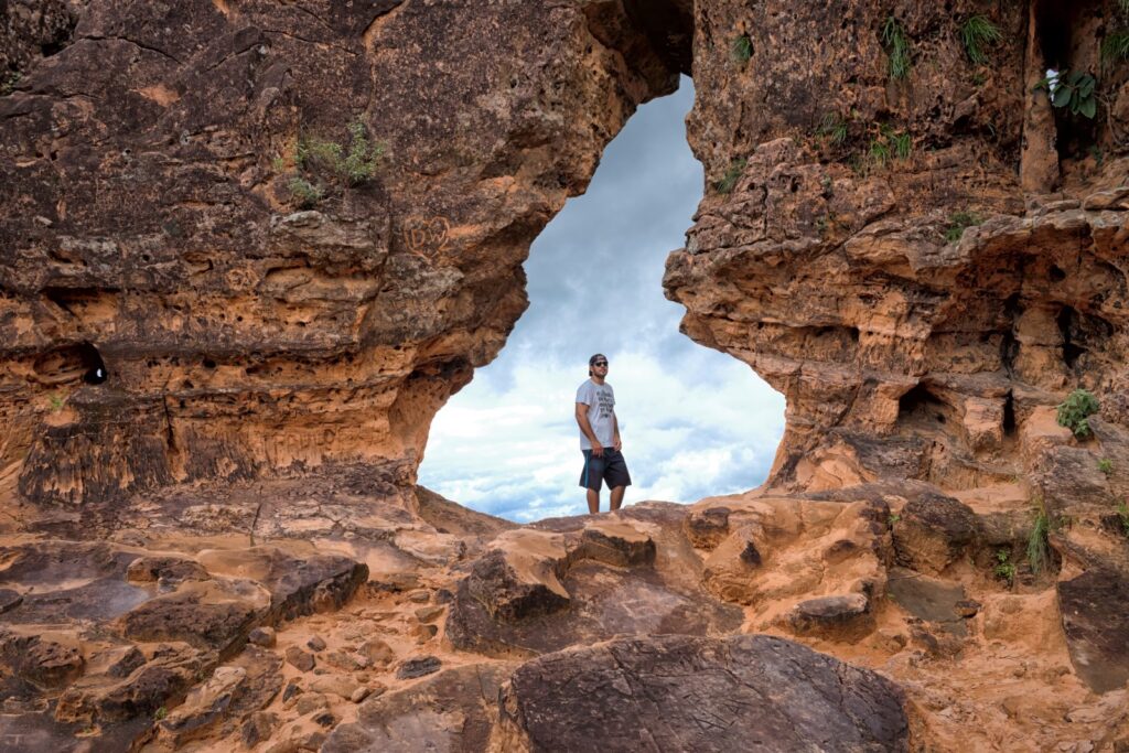 Visitante de pé dentro do arco de pedra do Portal da Chapada, formação de arenito com céu e nuvens ao fundo