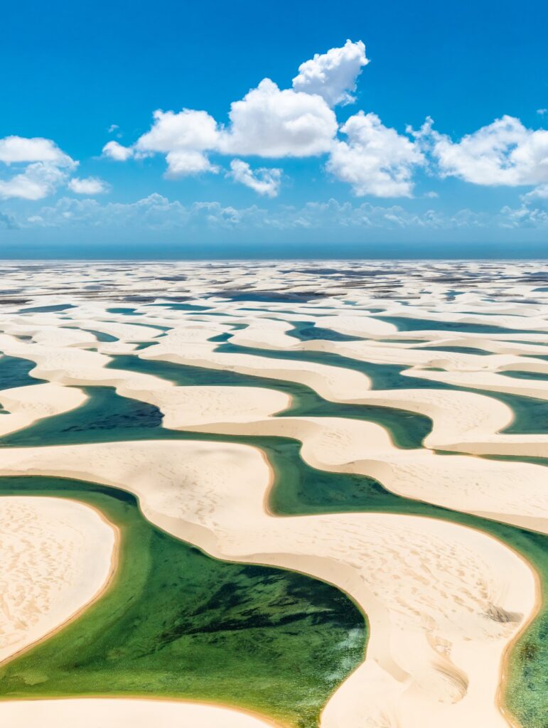 Vista aérea das dunas brancas dos Lençóis Maranhenses com lagoas verdes entre elas e céu azul com nuvens