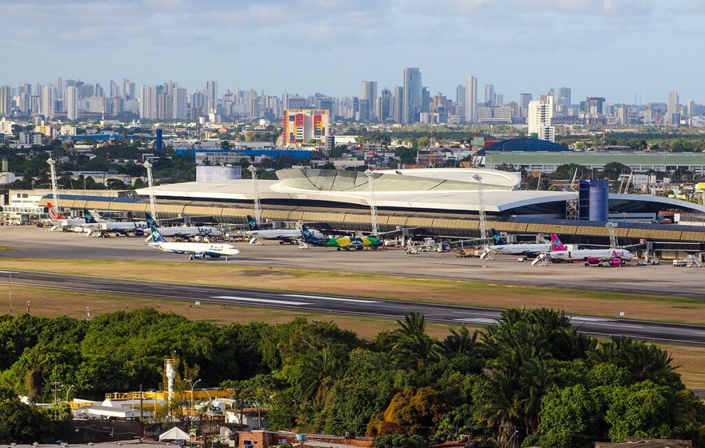 Vista aérea do Aeroporto Internacional do Recife, com aviões no pátio e os prédios da cidade ao fundo