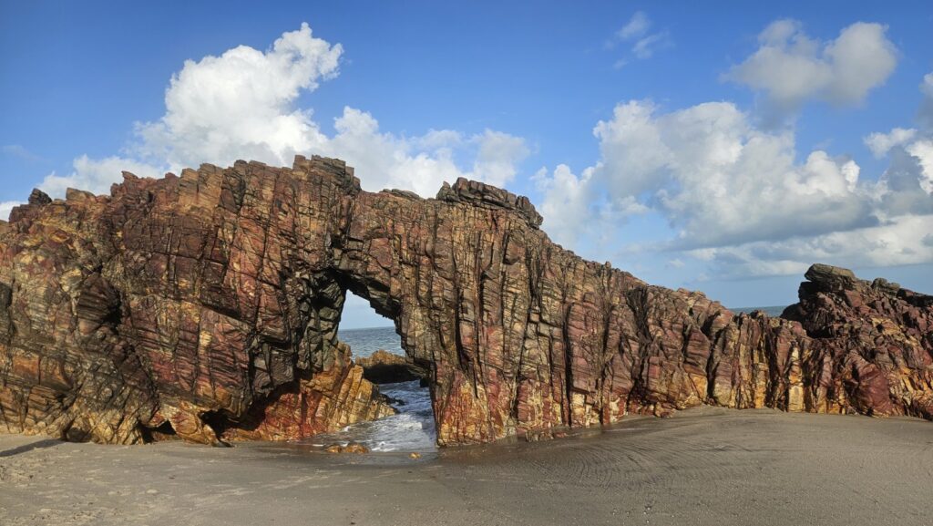 Pedra Furada de Jericoacoara, arco de rocha avermelhada sobre a areia, com céu azul e nuvens