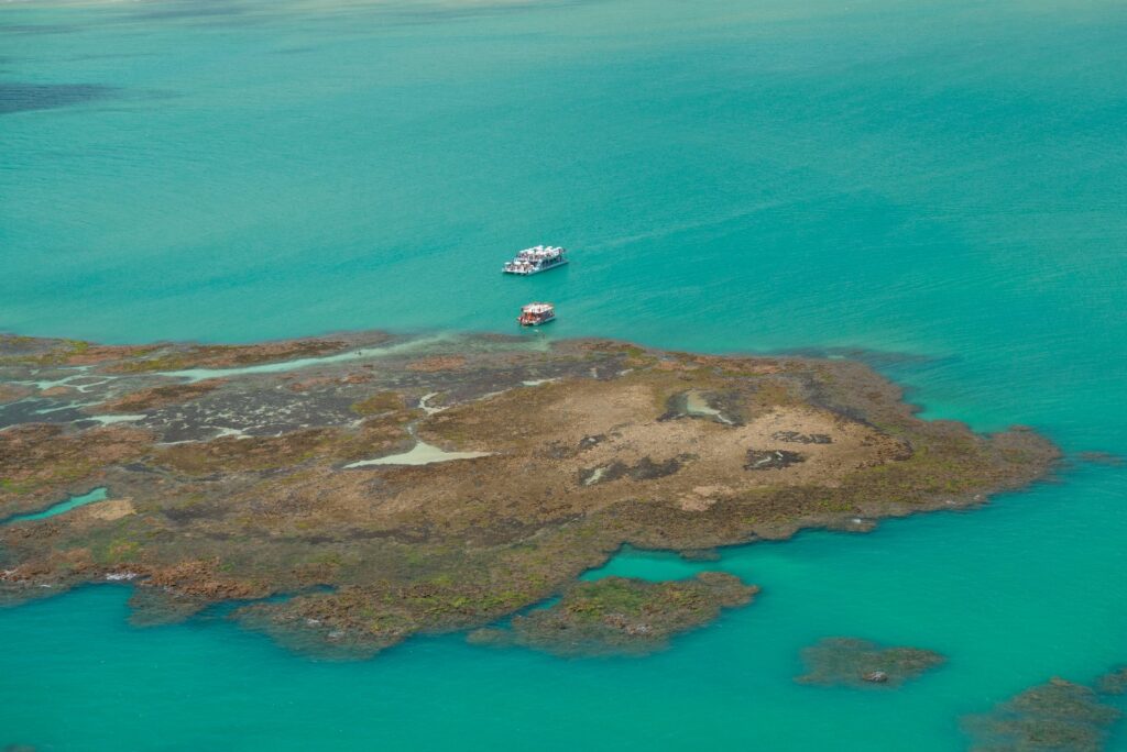 Vista aérea das piscinas naturais de Picãozinho na maré baixa, com recifes expostos e catamarãs ancorados no mar verde-claro