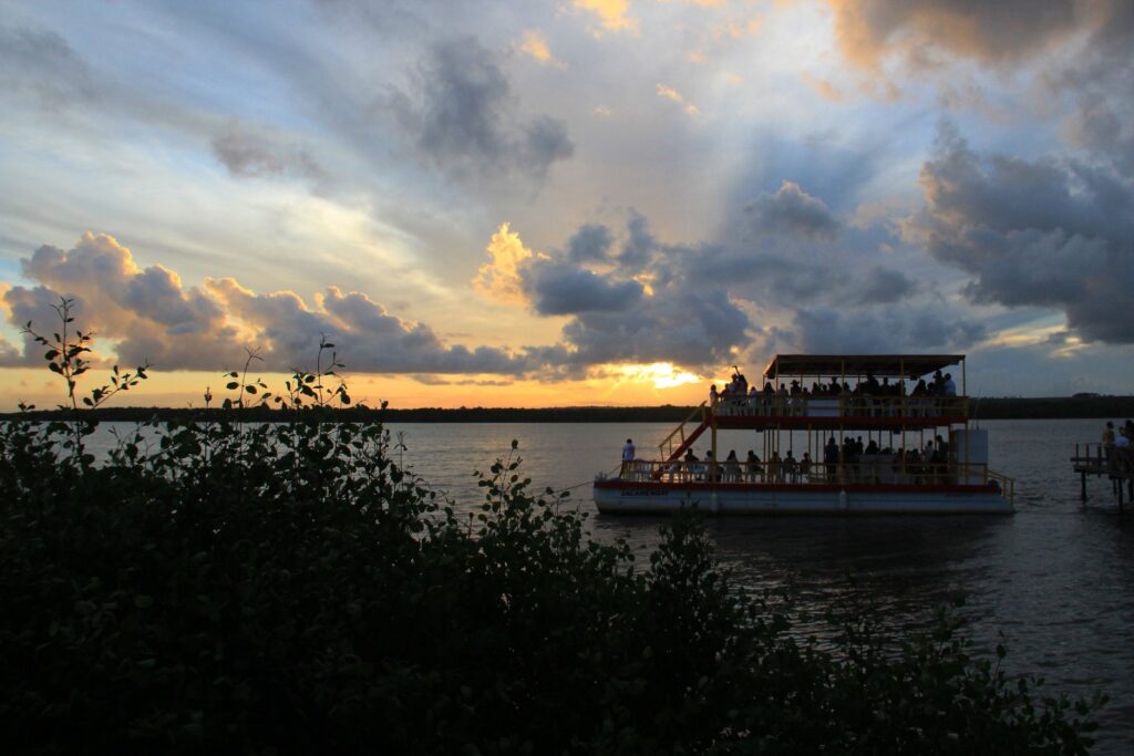 Pôr do sol sobre o rio Paraíba na Praia do Jacaré, em Cabedelo, com um barco de passeio iluminado e nuvens douradas