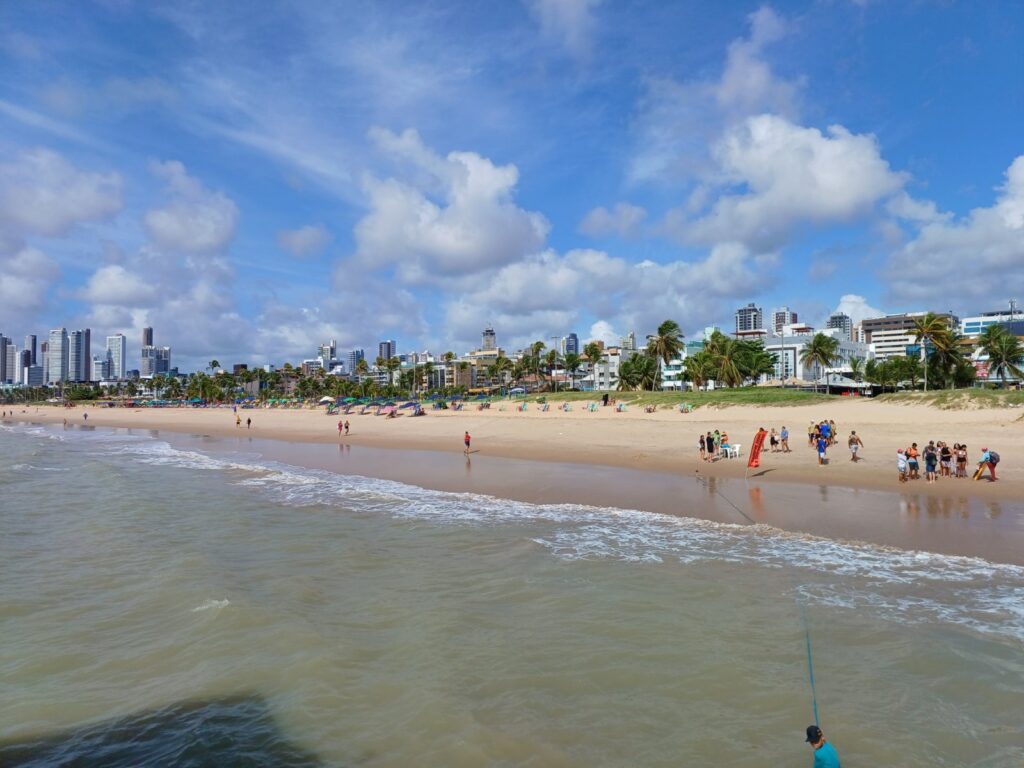 Praia de Tambaú, em João Pessoa, com faixa de areia, banhistas, coqueiros e prédios baixos da orla sob céu azul
