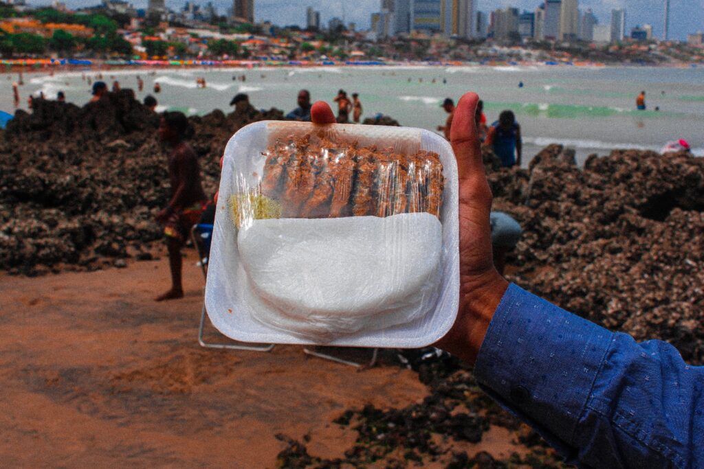 Mão segurando uma porção de ginga com tapioca, peixinhos fritos sobre tapioca, na praia de Ponta Negra