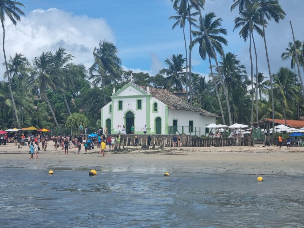 Capela de São Benedito, pequena igreja branca à beira-mar na Praia dos Carneiros, cercada de coqueiros e visitantes