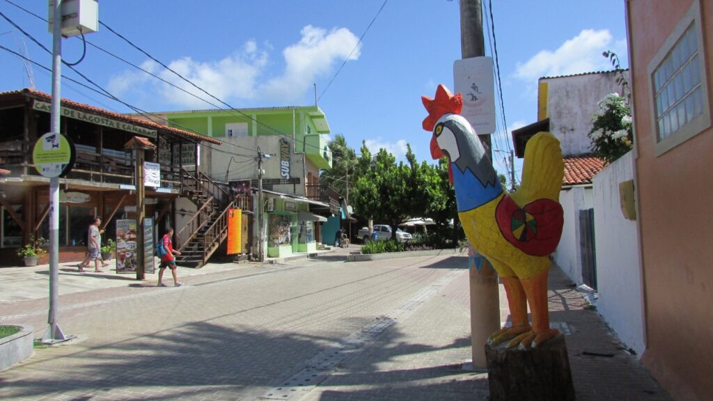 Rua da vila de Porto de Galinhas com uma grande escultura colorida de galinha em madeira na calçada