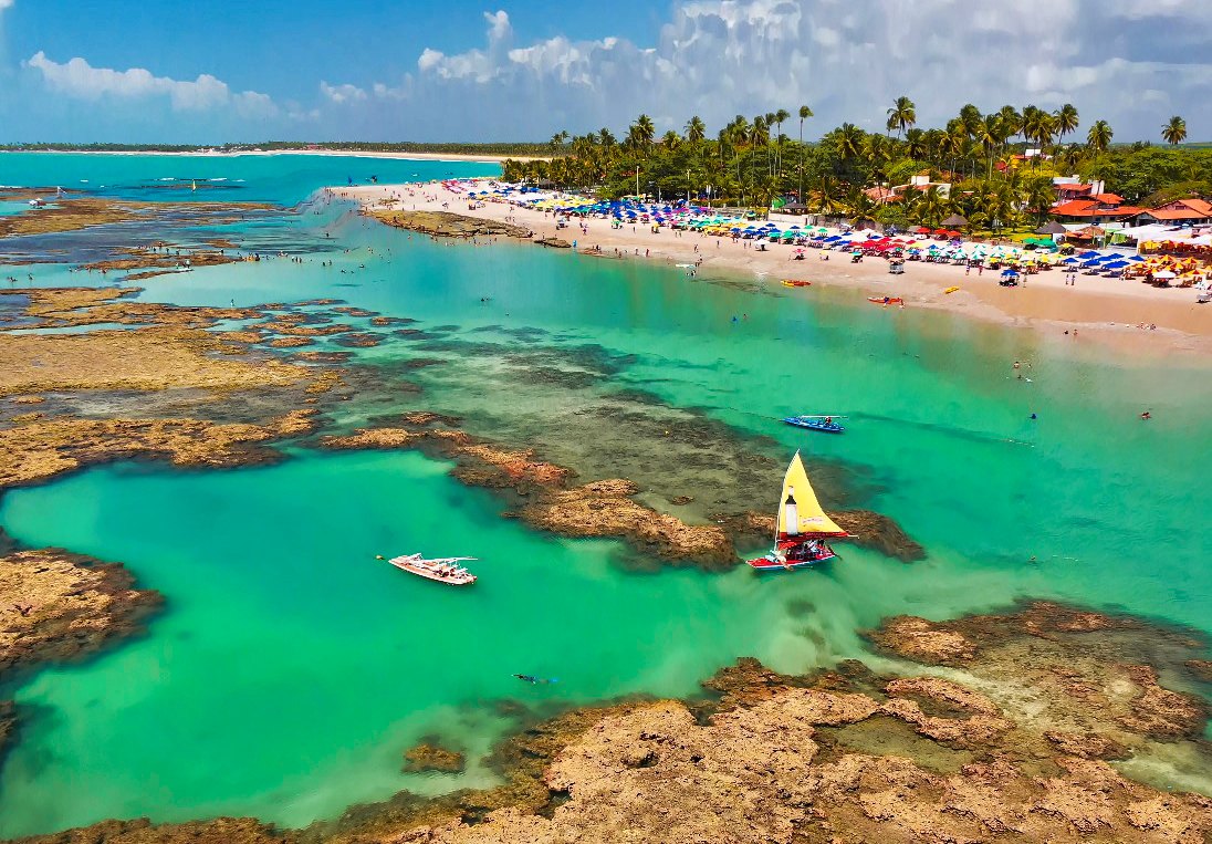 Vista aérea das piscinas naturais de Porto de Galinhas, com recifes, água azul-turquesa, jangadas coloridas e a praia cheia de guarda-sóis ao fundo