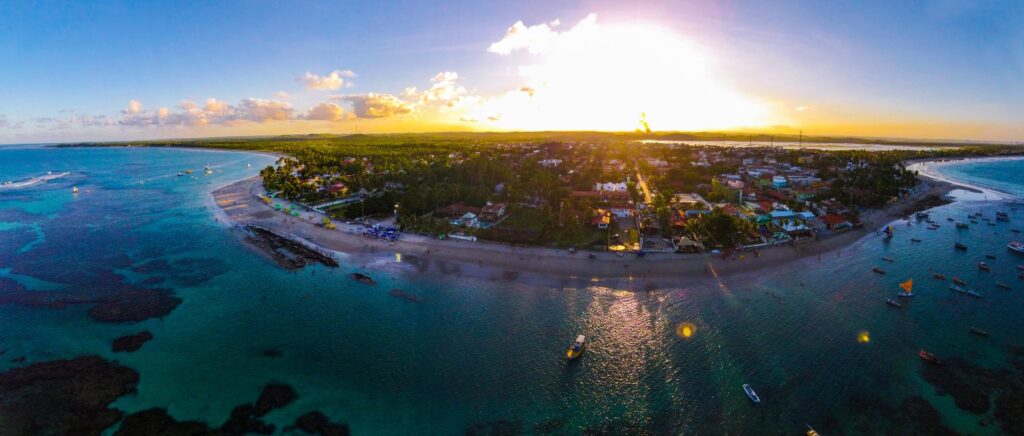 Vista aérea da vila de Porto de Galinhas ao entardecer, com a ponta de areia, os recifes e barcos ancorados no mar