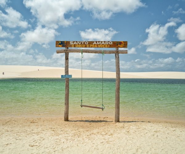 Balanço com placa de Santo Amaro à beira de uma lagoa de água verde entre as dunas dos Lençóis Maranhenses