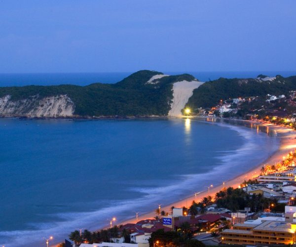 Orla de Ponta Negra ao entardecer, com as luzes dos hotéis acesas e o Morro do Careca ao fundo