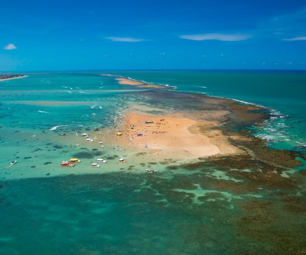 Vista aérea do banco de areia de Areia Vermelha, em Cabedelo, exposto na maré baixa, cercado de recifes e mar verde-claro, com barcos ancorados