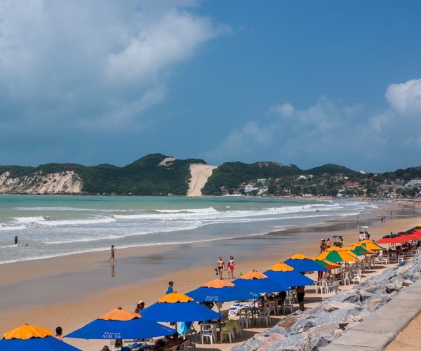Praia de Ponta Negra em dia de sol, com guarda-sóis coloridos na areia, mar calmo e o Morro do Careca ao fundo