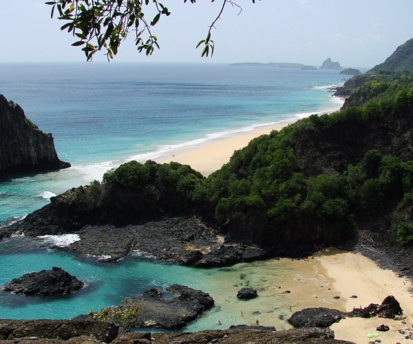 Baía dos Porcos com piscinas de pedra, uma longa faixa de areia ao fundo, o Morro Dois Irmãos à esquerda e o Morro do Pico no horizonte