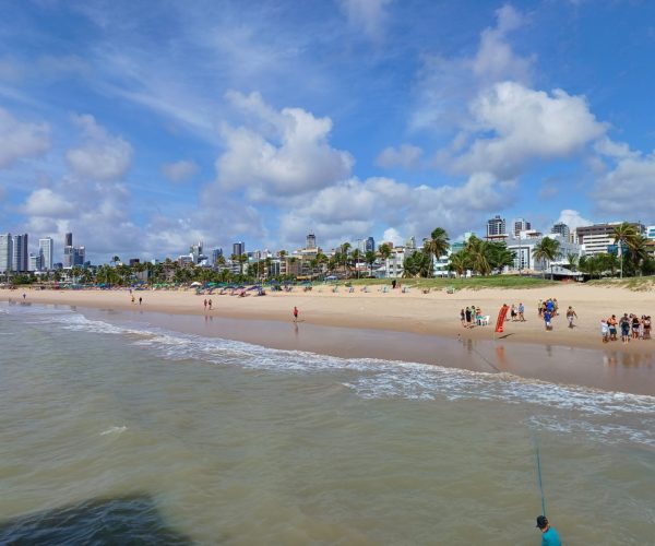 Praia de Tambaú, em João Pessoa, com faixa de areia, banhistas, coqueiros e prédios baixos da orla sob céu azul