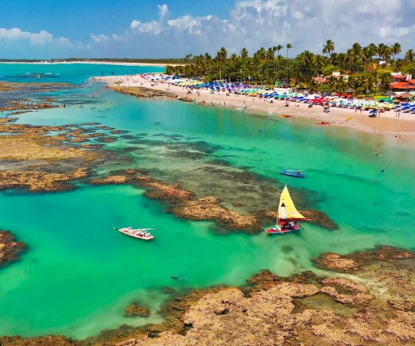 Vista aérea das piscinas naturais de Porto de Galinhas, com recifes, água azul-turquesa, jangadas coloridas e a praia cheia de guarda-sóis ao fundo